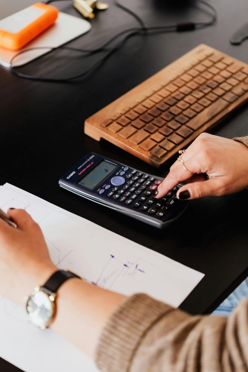 A person doing business analytics jobs role by typing into a black calculator and copying it on paper.